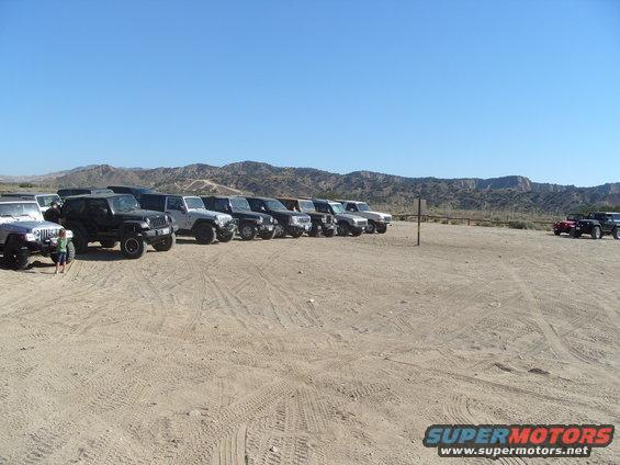 Multiple SUVs and trucks parked in a dirt lot with hills in the background
