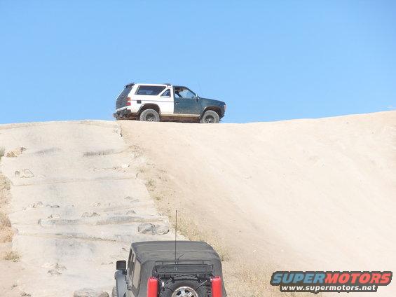 Two SUVs driving up a sandy hill, one white and green, the other black