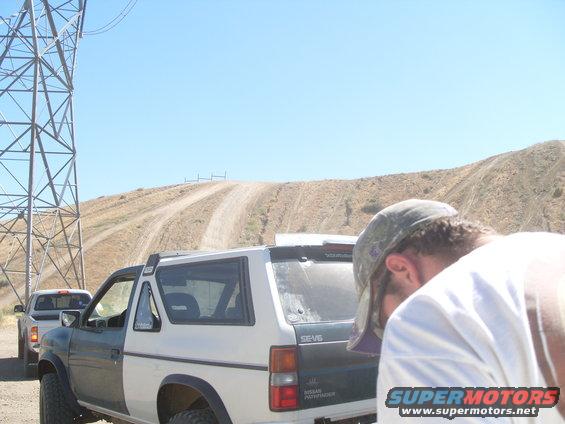 Two-tone Nissan Pathfinder SUV, rear view, with a white pickup truck behind it on a dirt road