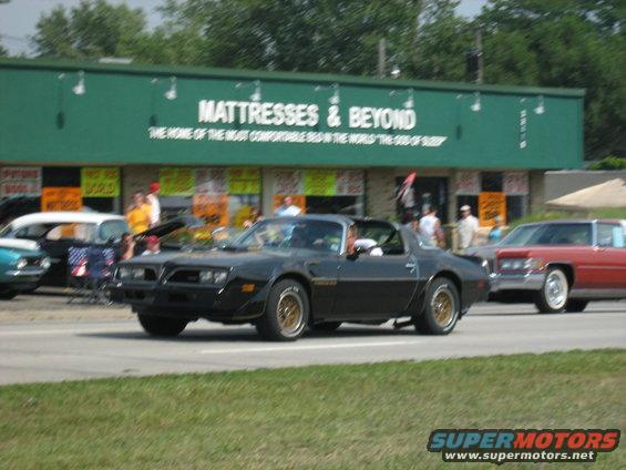 Black 1977 Pontiac Firebird Trans Am with gold wheels driving past a Mattress & Beyond store