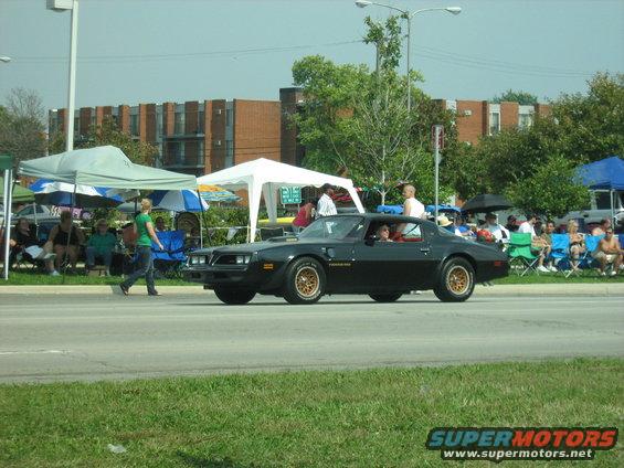 Black 1977 Pontiac Firebird Trans Am with gold wheels driving past spectators