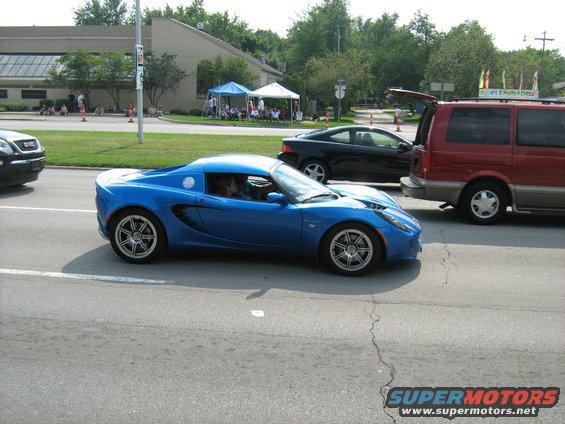Blue sports car with a driver, seen from the side on a street with other vehicles and tents in the background