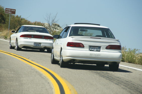 Two white sedans on a road, one with a "SCENIC VISTA 1/4 MILE" sign behind it. The rear car has a license plate reading "3PTF369" and the badge "PHE"