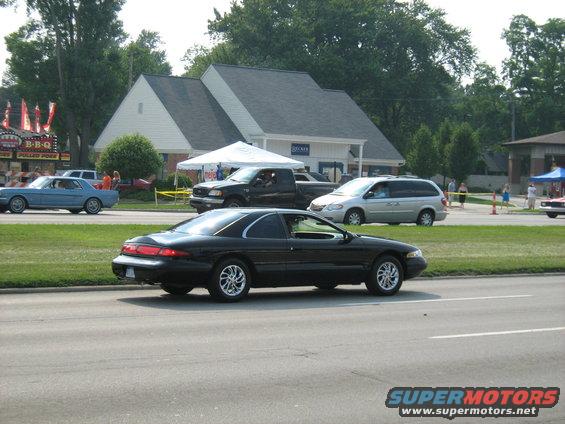 Black coupe driving past a building with tents and other cars on the road