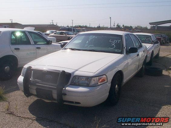White Ford Crown Victoria police car with a push bumper and 'LOT 3' written on the windshield. Another similar car is parked next to it