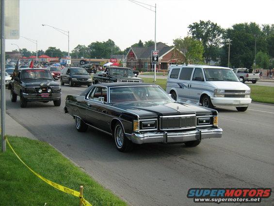 Black 1970s Mercury Grand Marquis coupe driving on a street, front view. Chrome grille and bumper, red interior visible