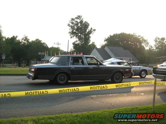 Dark sedan with a vinyl roof, another car behind it, and yellow caution tape in the foreground