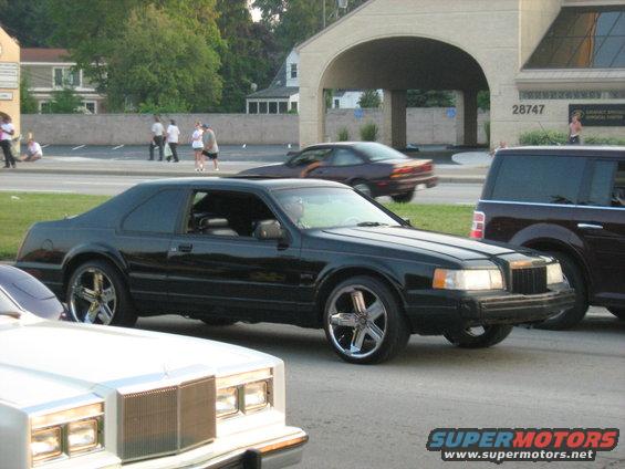 Black coupe with aftermarket wheels parked on a street, side view. A white car is in the foreground