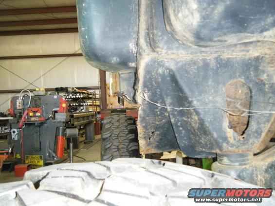 Close-up of a dark-colored vehicle frame and a large tire, with industrial machinery in the background