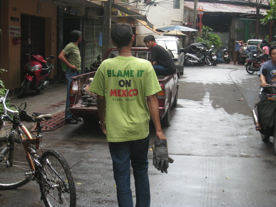Street scene with people, a red scooter, a pickup truck, bicycles, and motorcycles. Man in foreground wears a t-shirt that says 'BLAME IT ON MEXICO'