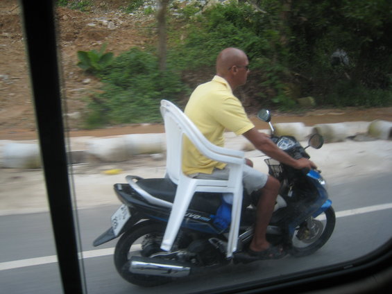 Man on a motorcycle with a white plastic chair strapped to the rear seat