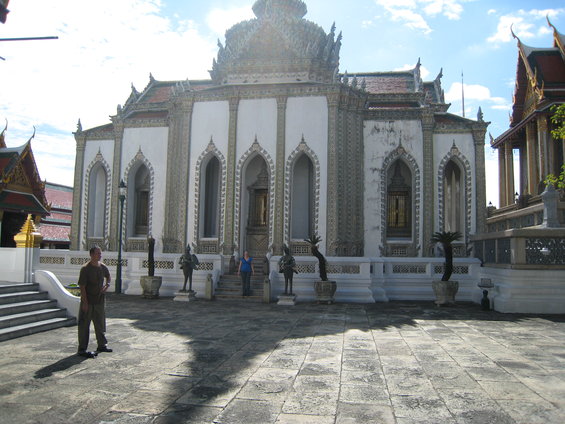 Man standing on stone courtyard in front of ornate white building with arched windows and decorative trim. Two people stand near entrance
