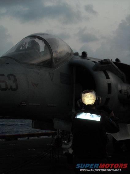 Military jet with number 63 on fuselage, on an aircraft carrier deck at dusk. A person in a reflective vest stands nearby