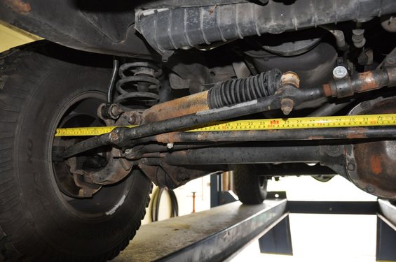 Undercarriage of a vehicle showing steering linkage and a yellow tape measure. Rusty components are visible