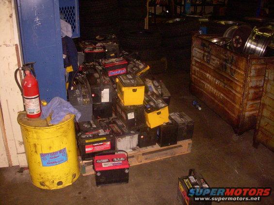 pile of car batteries on a pallet, with a fire extinguisher on a yellow drum nearby