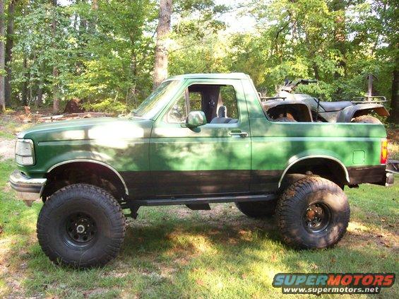 Lifted green Ford Bronco with black trim, an ATV in the bed, parked in a grassy, wooded area