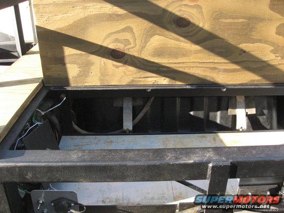 Open storage compartment on a trailer with wooden lid, showing wiring and metal supports
