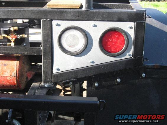 Close-up of two round tail lights, one clear and one red, mounted on a metal bracket. Trailer hitch visible below