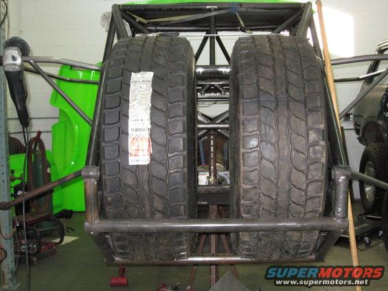 Two large off-road tires mounted on a custom frame, likely for a desert racing vehicle. A green body panel is visible to the left