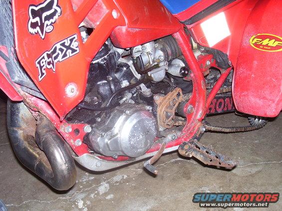 Close-up of a red Honda dirt bike engine, carburetor, exhaust pipe, and footpeg. Fox logo visible on the fairing