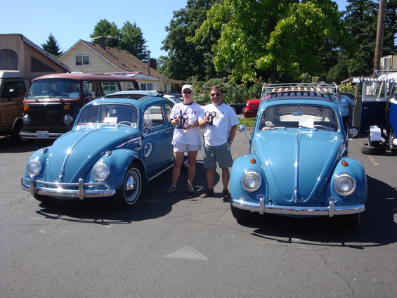 Two blue Volkswagen Beetles parked side-by-side, with people holding trophies