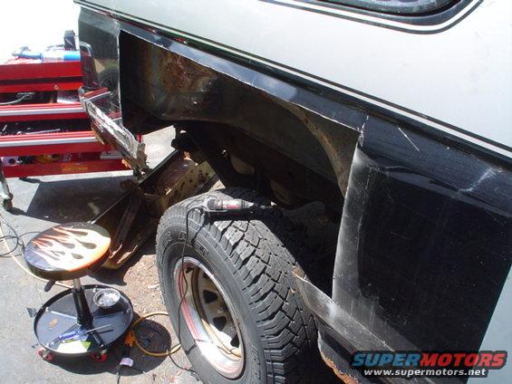 close-up of rusted rear quarter panel of a vehicle being worked on, with a tire and tool cart visible