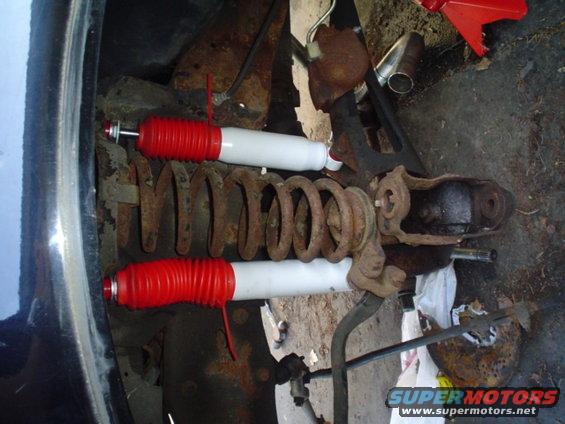 Rusty coil springs and white shocks with red bellows are mounted to a vehicle's suspension assembly