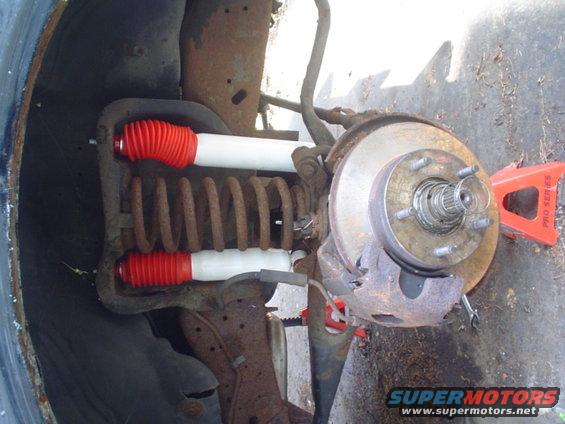 close-up of vehicle's front suspension with new red-booted shocks and rusty coil spring, brake rotor visible