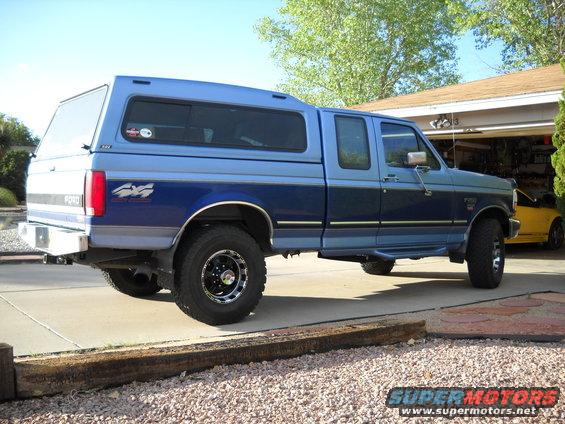 blue Ford pickup truck with camper shell, parked in driveway. Yellow car in garage
