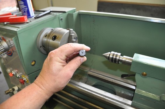 Hand holding a small cylindrical metal part in front of a metal lathe