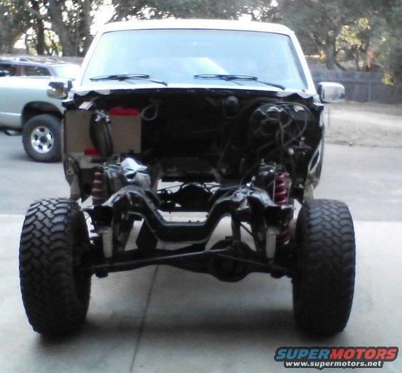 Truck chassis with front suspension and large tires, hood removed, showing engine bay components