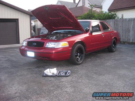 Red sedan, possibly a 2000 Ford Crown Victoria, parked outside with the hood open. Tools and rags are on the ground