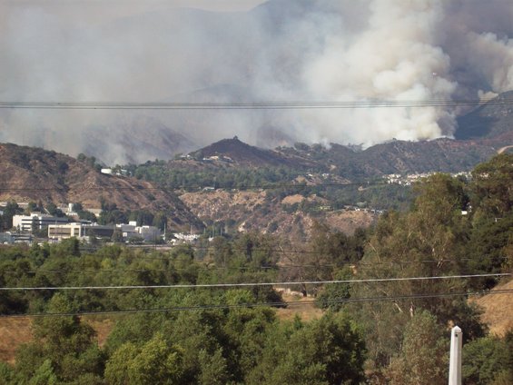 Smoke billows from a wildfire on a dry, hilly landscape with scattered buildings and trees