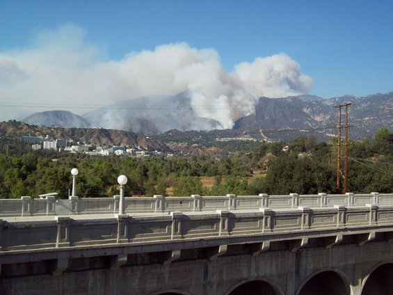 Smoke billows from a wildfire on a mountainside, with buildings visible in the mid-ground and a bridge in the foreground