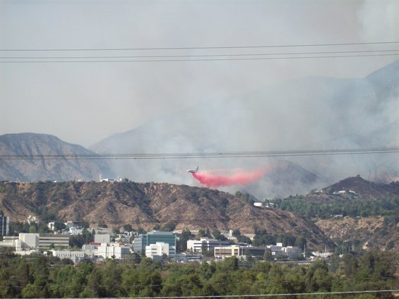 Firefighting aircraft drops red retardant over a hillside with buildings below