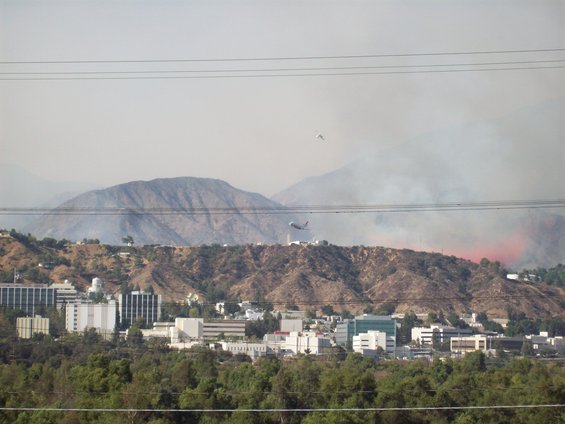 Wildfire burning on a hillside with an air tanker dropping water, buildings and trees in the foreground
