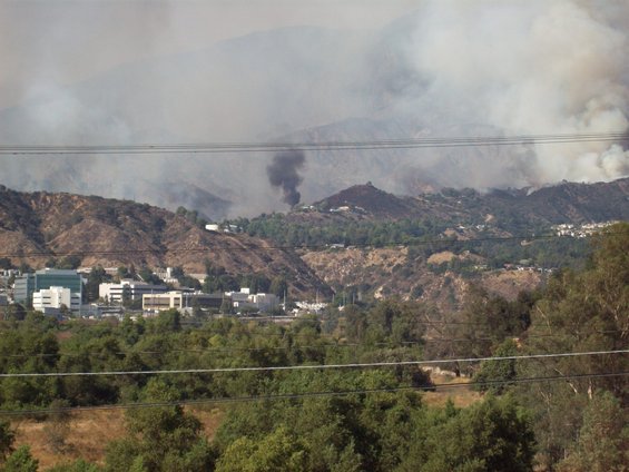 Smoke plumes rise from a wildfire in the hills above buildings and trees