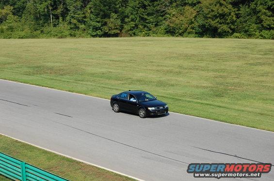 Black sedan with racing numbers on the window drives on a paved track with grassy fields and trees in the background