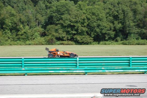 Orange track car with a driver wearing a helmet, on a race track with a green barrier in the foreground