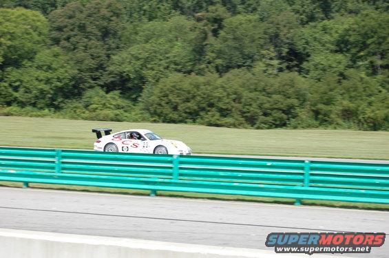 White Porsche 911 race car with a spoiler on a track, green guardrail in foreground