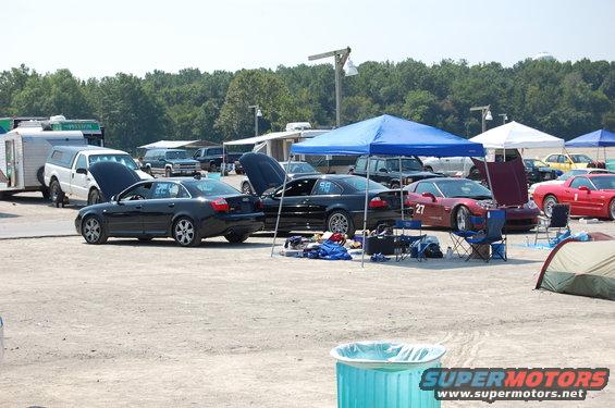 Race track paddock scene with black Audi, black BMW, red Corvette, and other vehicles under tents