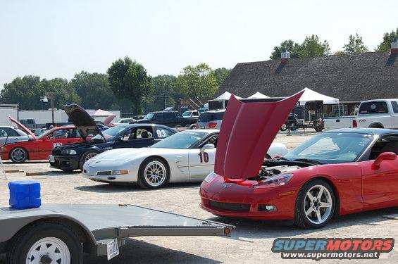 Several cars, including a red Corvette with its hood up, a silver Corvette with number 10, and a black BMW with number 93, parked at an outdoor event