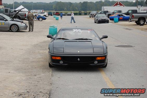 Front view of a black Ferrari F355 with racing number 355 on windshield. Other cars and tents in background