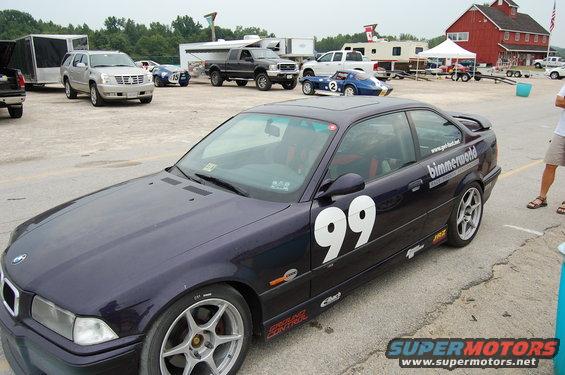 Dark purple BMW coupe with racing number 99 on the door, parked at a track with other vehicles and buildings in the background