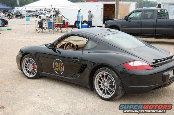 Black Porsche Cayman with racing number 26 on the door, parked on gravel. Other vehicles and tents are in the background
