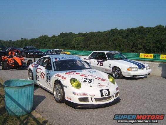 White Porsche 911 GT3 Cup car with yellow headlights and a white Ford Mustang with blue stripes on a racetrack