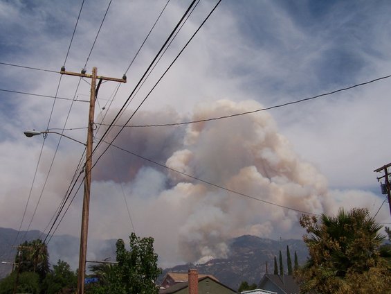 Large plume of smoke from a wildfire rises behind hills and houses, with power lines in the foreground