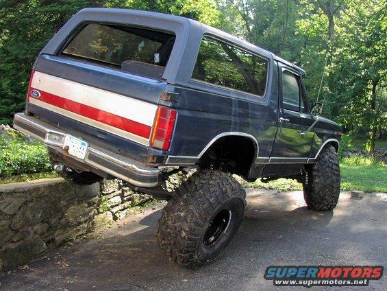 Dark blue Ford Bronco with red and white stripe on the side, lifted with big off-road tires, partially on a stone wall
