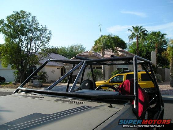 Interior view of a custom off-road vehicle with a roll cage and a red cloth draped over the passenger seat. A yellow SUV is visible in the background