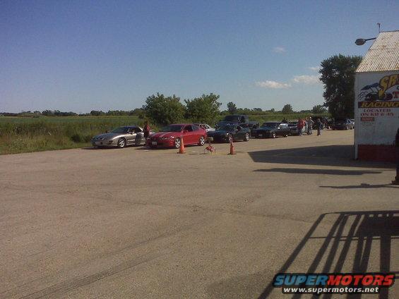 Several cars parked in a lot near a building with racing graphics; a field is in the background